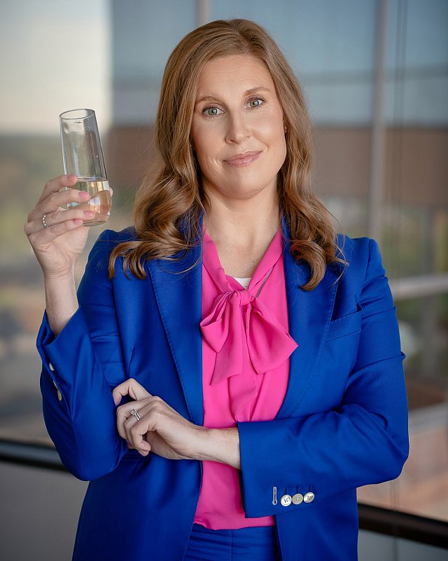 Photo of a female executive sales training coach in a blue jacket with a pink blouse, holding a glass of champagne. 